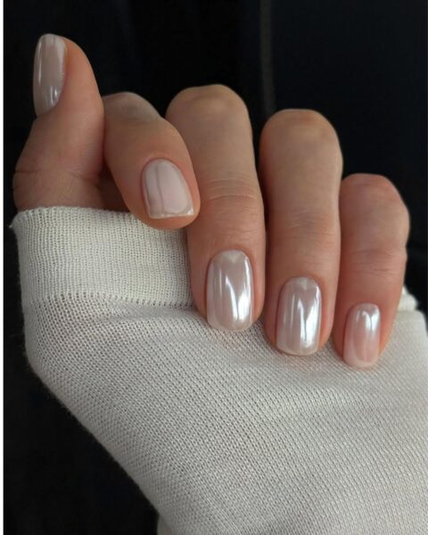 Zdjęcie dłoni z krótkimi, kwadratowymi paznokciami w kolorze białym z efektem mirror nails. Dłoń pozuje tak, by prezentować manicure. Widać fragment rękawa białego swetra.