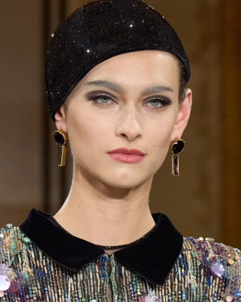 Zdjęcie portretowe modelki z pokazu Armani Prive. Ma czarną czapkę z cekinami na głowie, złote kolczyki z czarnymi kamieniami. Makijaż z nude ustami i cieniowanem oczu w stylu lat 50. Ubrana jest w kolorową bluzkę z czarnym kołnierzem.