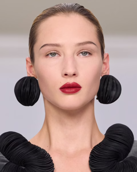 Zdjęcie modelki z pokazu Chocheng HC ze zbliżeniem na jej twarz z delikatnym makijażem i ustami w kolorze winnym. Ma blond, gładko związane włosy. W uszach masywne, czarne kolczyki i ubrana jest w czarną bluzkę z plisowanymi bufkami przy dekolcie.