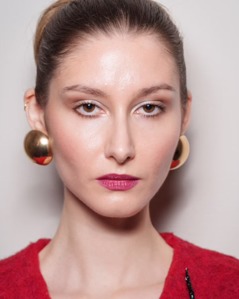 Zdjęcie portretowe modelki z pokazu Imane Ayissi HC. Ma blond włosy związane gładko w kok. W uszach obłe, złote kolczyki. Jest ubrana w czerwony sweter. Ma delikatny makijaż z borówkowymi ustami.