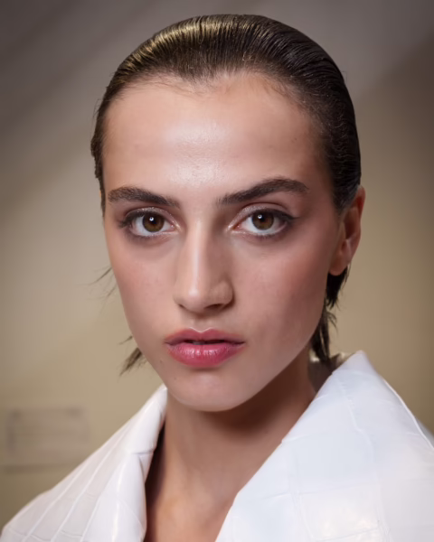 Zdjęcie portretowe modelki z pokazu Tamara Ralph HC. Ma ciemne włosy gładko zaczesane do tyłu. Ma delikatny makijaż i usta pomalowane satynową pomadką, bo to modny makijaż ust na jesień 2025. Jest ubrana w białą marynarkę.