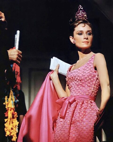 Opis alt dla zdjęciaNa zdjęciu widać aktorkę Audrey Hepburn w zbliżeniu od klatki piersiowej w górę. Ma na sobie bogato zdobioną, różową suknię bez rękawów z błyszczącymi, łuskowatymi zdobieniami i satynową kokardą w talii. Na głowie ma tiarę. Trzyma w ręce kawałek różowego materiału. Zdjęcie zostało zrobione w słabo oświetlonym pomieszczeniu, co nadaje mu tajemniczy, filmowy klimat. Kreacja pochodzi z filmu "Jak ukraść milion dolarów" z 1966 roku.