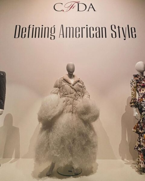 Na zdjęciu widać fragment wystawy mody. Na białej ścianie nad manekinami widnieje napis: "CFDA Defining American Style". W centrum, na podeście, stoi manekin w obszernej, białej kurtce przypominającej puchową, z bardzo dużymi, puszystymi rękawami i dołem. To kreacja, która wyróżnia się na tle pozostałych. Po lewej i prawej stronie stoją manekiny w innych strojach: po lewej w błyszczącym, szarym kombinezonie, a po prawej w długiej, wielowarstwowej sukience z kwiecistym wzorem.