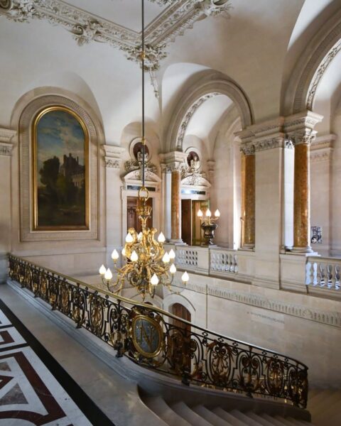 Zdjęcie przedstawia bogato zdobione wnętrze Luwru (Musée du Louvre) w Paryżu. W centralnej części widać reprezentacyjną klatkę schodową z kamiennymi stopniami i kutą, czarno-złotą balustradą. Nad schodami wisi okazały, złoty żyrandol w stylu empire. Na ścianach widać białe sztukaterie i rzeźbione detale, a w oświetlonej niszy po lewej stronie znajduje się duży obraz w złoconej ramie z pejzażem. Marmurowa podłoga i kolumny podkreślają klasyczny, królewski charakter wnętrza.