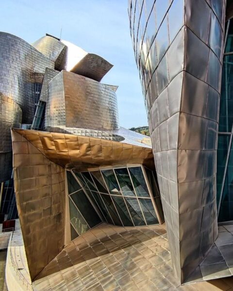 Zbliżenie na dynamiczną, futurystyczną fasadę Muzeum Guggenheima w Bilbao, zaprojektowanego przez Franka Gehry'ego. Powierzchnia budynku jest pokryta tytanowymi płytami, które tworzą organiczne, falujące kształty, odbijające światło słoneczne. Struktura łączy niemożliwe kąty, zakrzywione ściany i szklane elementy, tworząc efekt abstrakcyjnej rzeźby na tle błękitnego nieba. Jest to symboliczne ujęcie nowoczesnego designu i innowacyjności w architekturze.
