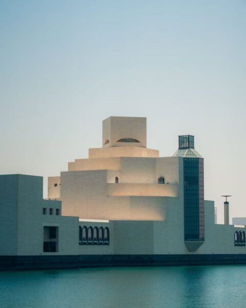 Zdjęcie przedstawia Muzeum Sztuki Islamskiej (Museum of Islamic Art, MIA) w Dosze w Katarze, zaprojektowane przez I.M. Pei. Budynek o jasnopiaskowym kolorze jest zbudowany z geometrycznych brył, które wznoszą się kaskadowo, tworząc wrażenie masywnej fortecy na wodzie. Architektura łączy islamskie tradycje z minimalistycznym, nowoczesnym stylem. Na pierwszym planie widoczna jest turkusowa woda Zatoki Perskiej, nad którą wznosi się budowla. Całość jest skąpana w jasnym, lekko zamglonym świetle słonecznym.