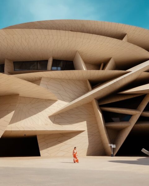 Zdjęcie przedstawia dynamiczną fasadę Muzeum Narodowego Kataru (National Museum of Qatar) w Dosze, która naśladuje kształt róży pustyni. Beżowa, wielowarstwowa struktura o nieregularnych geometrycznych płaszczyznach jest oświetlona jasnym słońcem, kontrastując z intensywnie błękitnym niebem. Na pierwszym planie, u podstawy monumentalnego budynku, stoi jedna kobieta ubrana w długą, pomarańczowo-białą sukienkę o wyrazistym, geometrycznym wzorze, która dodaje scenie koloru i perspektywy.