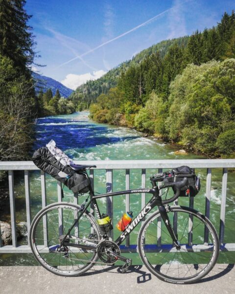 Zdjęcie krajobrazu mijanego po trasie rowerowej Alpe Adria Radweg. Widać most, na którym stoi rower oparty o barierkę. W tle widać rwącą rzekę oraz połacie lasu.