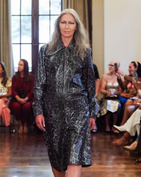 Zdjęcie podczas São Paulo Fashion Week z pokazu Patricia Viera. Po wybiegu idzie dojrzała modelka z długimi, siwymi włosami. Ubrana jest w kurtkę i spódnicę w kolorze czarnym z motywem panterki.