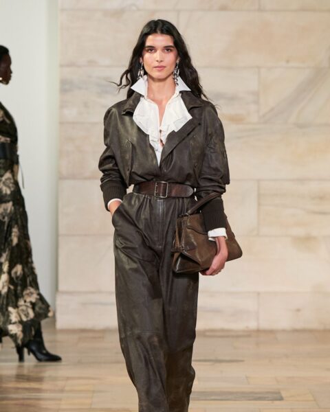 Zdjęcie podczas New York Fashion Week z pokazu Ralpha Laurena. Modelka, ciemnowłosa, idzie na wybiegu prezentując białą koszulę, kombinezon w kolorze khaki i ma pod ręką brązową torebkę.