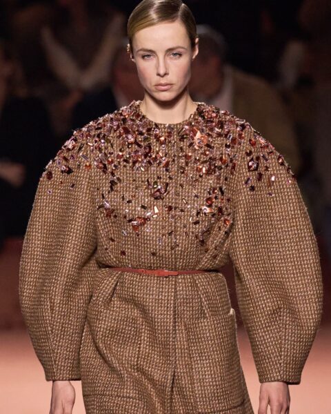 Zdjęcie podczas Milan Fashion Week z pokazu Fendi. Widać modelkę ubraną w brązową sukienkę w kratkę z szerokimi rękawami, paskiem w talii oraz zdobieniem przy kołnierzu.
