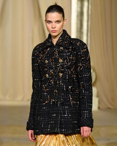 Zdjęcie podczas Paris Fashion Week z pokazu Chanel. Widać modelkę ubraną w czarną, tweedową marynarkę ze złotymi zdobieniami oraz złotą spódnicę.