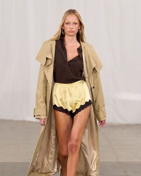 Zdjęcie podczas Copenhagen Fashion Week z pokazu Rotate. Widać modelkę, blondynkę w maślanych szortach z koronką, brązowym kardiganem i długim płaszczem trenczem.
