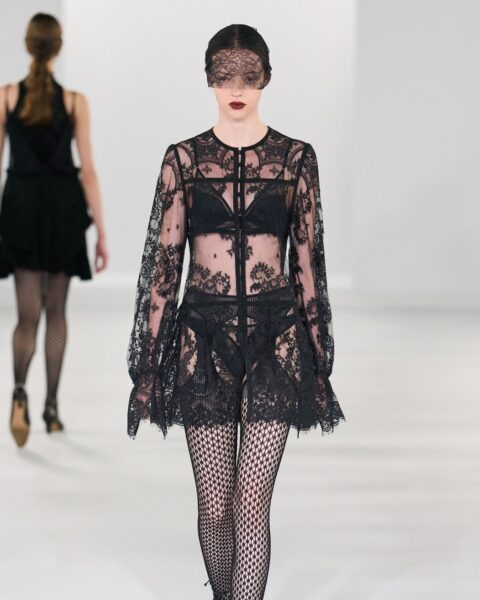 Zdjęcie podczas Tokyo Fashion Week z pokazu Flabelus. Po wybiegu idzie modelka ubrana w koronkową sukienkę mini, kabaretki rajstopy oraz koronkową woaką na twarzy.