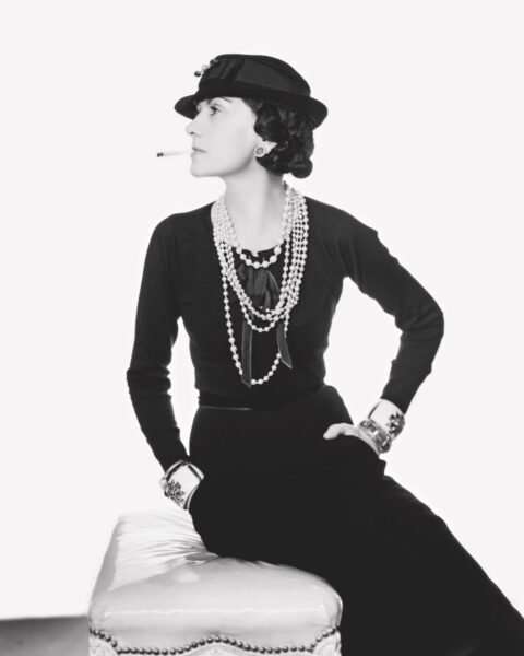Największe ikony stylu: zdjęcie Coco Chanel. Jest w sukience typu mała czarna, ma na sobie sznur pereł, kapelusz na głowie. Pozuje na pufie, ale obraca się w bok. Pali papierosa. Zdjęcie jest czarno-białe.