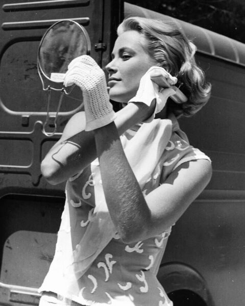 Zdjęcie Grace Kelly pozującej na tle dużego samochodu. Jest ubrana w bluzkę, spódnicę, na rękach ma białe rękawiczki. Włosy blond, falowane. Poprawia je, przeglądając się w lusterku.
