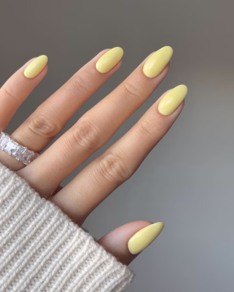 Zdjęcie dłoni z długimi, migdałowymi paznokciami. Są pomalowane na kolor maślanej żółci, czyli modny odcień butter yellow. Na palcu serdecznym widać srebrną obrączkę z kamieniami.