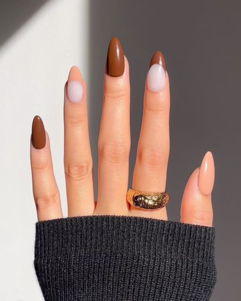 Zdjęcie dłoni z długimi, migdałowymi paznokciami. Są częściowo pomalowane na jednolity kolor brązowy, a częściowo pomalowane w stylu french manicure w kolorze brązowym. Wszystkie odcienie brązu mają ciepłe, czekoladowe tony. Na palcu serdecznym widać masywny, złoty pierścionek.