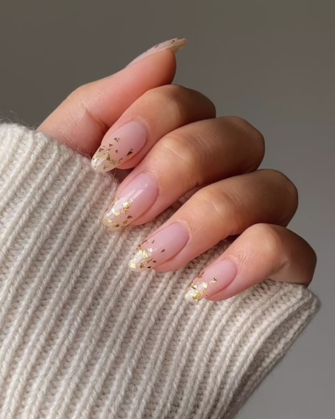 Zdjęcie dłoni z długimi, owalnymi paznokciami pomalowanymi na kolor nude i przy brzegach ozdobione złotymi płatkami folii. Dłoń pozuje tak, by prezentować manicure.