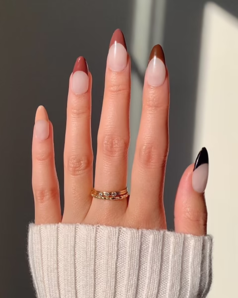 Zdjęcie dłoni ze zbliżeniem na długie, migdałowe paznokcie ze wzorem french manicure, ale zamiast białych końcówek są brązowe, na każdym paznokciu inny odcień. Na środkowym palcu widać złoty pierścionek.