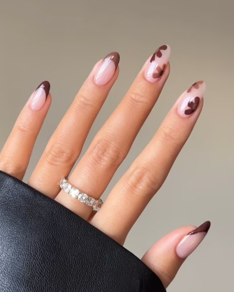 Zdjęcie dłoni z długimi, owalnymi paznokciami pomalowanymi na kolor nude i ozdobione wzorem liści w kolorze brązowym oraz brązowym french manicure. Na palcu środkowym widać pierścionek wysadzany kryształami. Widać fragment rękawa czarnej, skórzanej kurtki.