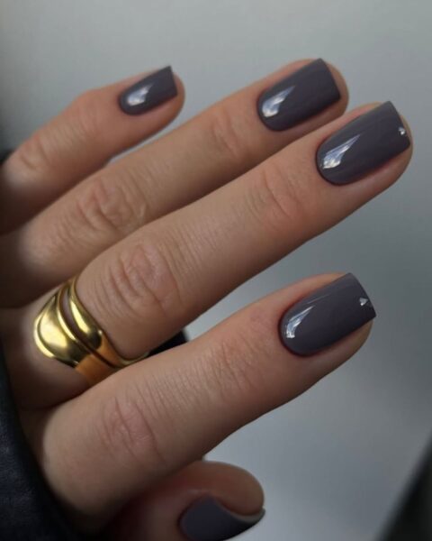 Elegancki manicure w odcieniu szarego lakieru. Krótkie paznokcie o kwadratowym kształcie, na serdecznym palcu mała, srebrna cyrkonia. Dłoń zdobi duży, złoty pierścionek o nieregularnym kształcie.