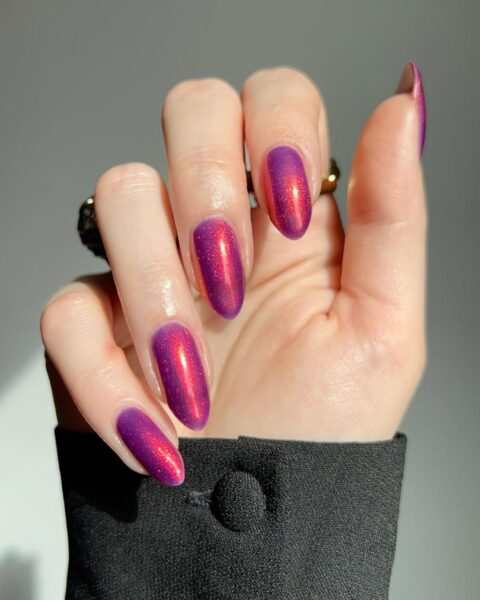 Lśniący, mieniący się manicure, który zmienia kolor z fioletowego na różowy, w zależności od kąta padania światła. Paznokcie o owalnym kształcie, a dłoń leży na czarnej koszuli z widocznym guzikiem. Na palcu serdecznym widać złoty pierścionek.