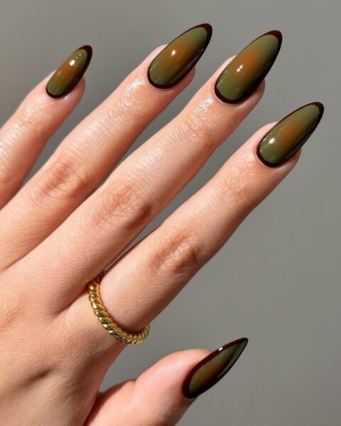 Długie, migdałowe paznokcie ze stylizacją w stylu "aura nails". Na oliwkowozielonej bazie widać brązowo-pomarańczową mgiełkę. Całość zdobi cienki, bordowy kontur. Dłoń ozdabia złoty, pleciony pierścionek.