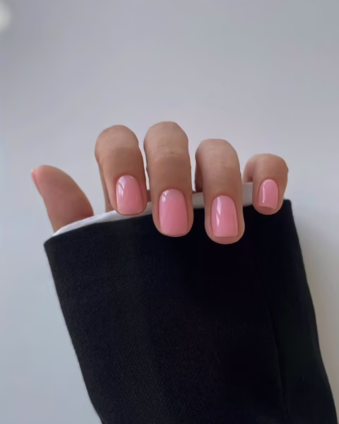 Zdjęcie dłoni z krótkimi, owalnymi paznokciami pomalowanymi na kolor jasnego różu. Dłoń pozuje tak, by prezentować manicure. Widać fragment rękawa białej koszuli schowanej pod czarnym rękawem marynarki.