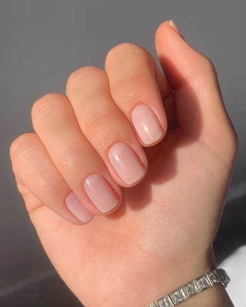 Zdjęcie dłoni ze zbliżeniem na paznokcie na spotkanie biznesowe z krótkimi, kwadratowymi płytkami pomalowanymi na kolor nude. Dłoń pozuje tak, by prezentować manicure. Na nadgarstku widać srebrną bransoletkę. 