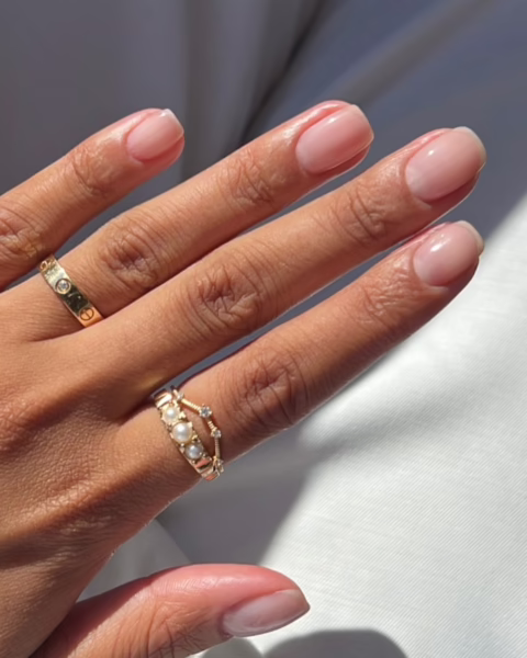 Zdjęcie dłoni ze zbliżeniem na biznesowy manicure: średniej długości paznokcie o kwadratowym kształcie w kolorze nude. Na palcach widać złote pierścionki. Dłoń pozuje na tle białych spodni.