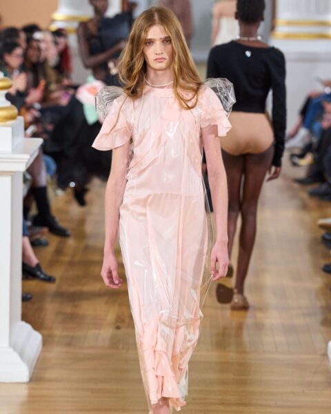 Awangardowy romantyzm z pokazu Simone Rocha: modelka w różowej sukience midi z falbanami, przykrytej warstwą przezroczystego plastiku. Inspiracja dla kobiet z klasą, ceniących artystyczną modę i unikalne faktury.