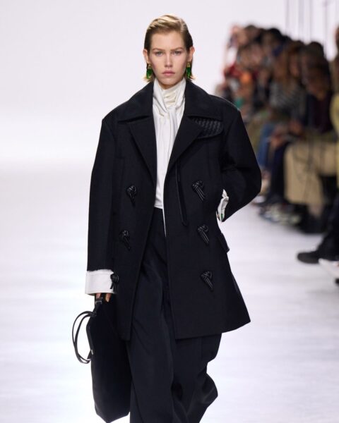 Rzeźbiarska elegancja i minimalizm z pokazu Bottega Veneta: modelka w czarnym, strukturalnym płaszczu o fasonie oversize, z luźnymi spodniami i białymi sneakersami. Inspiracja do budowania mody na klasę poprzez formę.