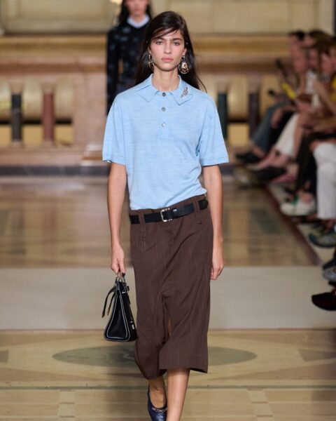 Stylizacja z pokazu Tory Burch S26. Modelka w eleganckiej, błękitnej koszulce polo i spódnicy midi w kolorze gorzkiej czekolady. Idealna inspiracja do stworzenia nowoczesnego, klasycznego looku w duchu preppy.