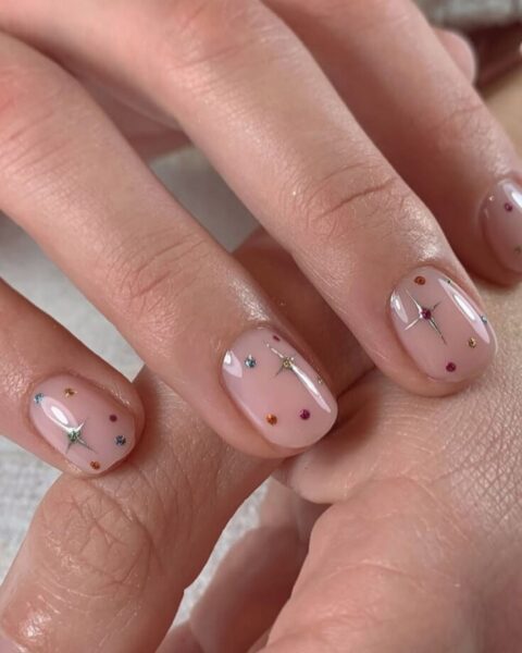 Zdjęcie dłoni z krótkimi, owalnymi paznokciami w kolorze nude i ozdobione motywem gwiazdek oraz kryształków. Dłonie pozują tak, by prezentować manicure.