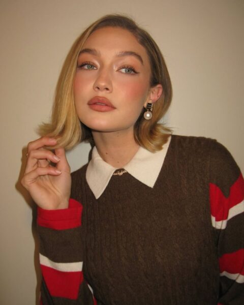 Modelka Gigi Hadid w stylizacji w klimacie preppy chic. Gigi ma na sobie brązowy sweter z kontrastowym kołnierzykiem i aplikuje modny makijaż, czyli kocie oko z różanym różem i ustami w odcieniu nude. Uwagę zwracają krótkie, pofalowane włosy w stylu boba i eleganckie kolczyki z perłami.