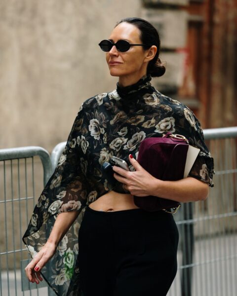 Elegancka kobieta w czarnych, wąskich okularach przeciwsłonecznych idzie ulicą, prezentując stylizację street fashion. Ma na sobie czarną, zwiewną bluzkę z florystycznym motywem w odcieniach beżu i zieleni, odsłaniającą delikatnie brzuch. Dół stanowią klasyczne, czarne spodnie z wysokim stanem. W dłoni trzyma burgundową torebkę typu clutch. Zdjęcie ilustruje minimalistyczny, ale wyrafinowany chic, charakterystyczny dla paryskiej elegancji.