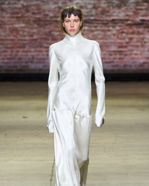 Zdjęcie modelki z pokazu marki Fforme. Ma na sobie białą, satynową sukienkę maxi z długimi rękawami i niską talią. Ma ciemne włosy, krótkie i z grzywką. Idzie po wybiegu.