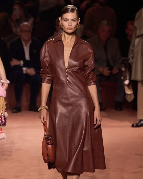Zdjęcie modelki z pokazu marki Fendi. Idzie po wybiegu, ma na sobie skórzaną, brązową sukienkę o rozkloszowanym dole, krótkimi rękawami, dopasowaną górą z dekoltem V. W ręce ma torebkę brązową. Włosy blond, związane gładko, a w uszach złote kolczyki.