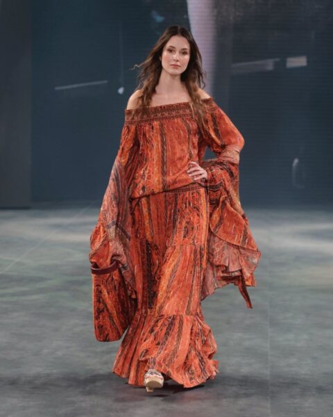 Modelka w zwiewnej, pomarańczowej sukience w stylu boho idzie po wybiegu. Sukienka ma dekolt odsłaniający ramiona, bufiaste rękawy i wielowarstwowy dół, a całość zdobi misterny wzór. Modelka ma długie, rozpuszczone włosy i delikatny makijaż. Tło jest ciemne i minimalistyczne, co skupia uwagę na stroju.