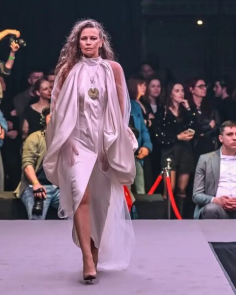 Modelka w długich, falowanych włosach idzie po wybiegu w błyszczącej, białej sukience. Kreacja ma asymetryczny dół i drapowany golf, który rozszerza się w szerokie, luźne rękawy z transparentnego materiału. Na szyi modelki wisi duży, złoty naszyjnik. W tle widać publiczność, a po lewej stronie fotografa z aparatem.