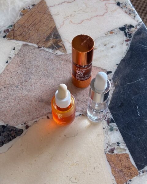 Ujęcie z góry trzech miniaturowych produktów do pielęgnacji skóry, ustawionych na eleganckim marmurowym blacie w ciepłych, brązowych i białych odcieniach. Produkty to: miedziana butelka Clarins, pomarańczowy olejek w szklanej butelce z pipetą oraz srebrna butelka serum. Kompaktowy zestaw esencjonalnych kosmetyków do podróży, skupiający się na wzmocnieniu bariery skórnej i głębokim nawilżeniu.