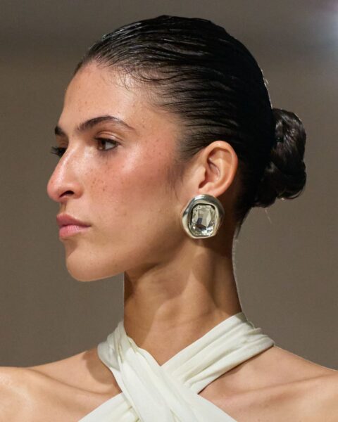 Zdjęcie przedstawia modelkę z bliska, na wybiegu PatBO, z fryzurą w typie slick back bun – włosy są gładko zaczesane do tyłu i upięte w kok. Na twarzy widać delikatny, świetlisty makijaż i naturalne piegi. Ma na sobie kremową sukienkę z marszczeniami przy szyi i kolczyki z dużym, błyszczącym kamieniem.