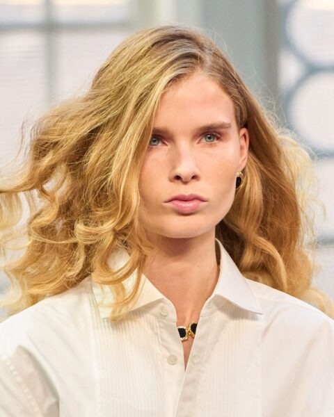 Zdjęcie przedstawia zbliżenie na twarz modelki z pokazu Mithridate. Ma jasne, długie, bujne i pofalowane włosy w stylu soft waves, które swobodnie opadają na ramiona. Ubrana jest w białą koszulę z zapięciem na guziki, pod szyją widać czarny naszyjnik, a w uchu ma mały, złoty kolczyk.