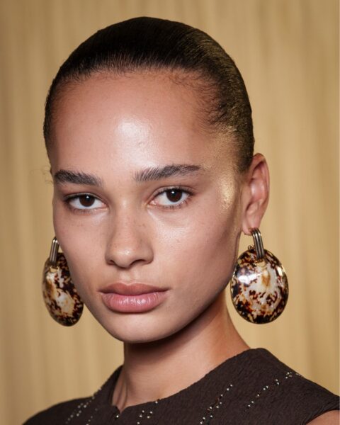 Zdjęcie przedstawia zbliżenie na twarz modelki z pokazu Tory Burch. Ma gładko zaczesane do tyłu włosy, które podkreślają makijaż typu no make-up. Na uszach ma duże, owalne kolczyki w brązowo-beżowym odcieniu z efektem muszli.