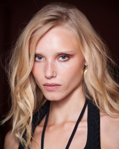 Zdjęcie przedstawia zbliżenie na twarz modelki zza kulis pokazu Marka Fast. Ma jasne, pofalowane włosy w odcieniu blond, swobodnie opadające na ramiona. Delikatny, lekko rozświetlony makijaż podkreśla naturalne rysy twarzy. Ma na sobie czarną sukienkę z ramiączkami wiązanymi na szyi.