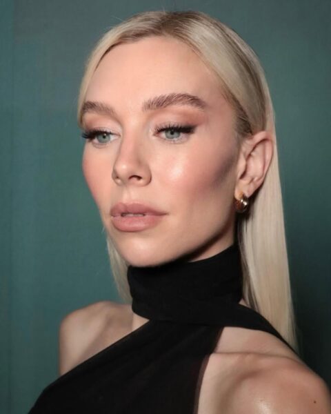 Portret Vanessy Kirby w eleganckim, czarnym stroju z golfem. Ma gładko zaczesane, platynowe blond włosy i makijaż z wyraźną, uniesioną kreską na powiece (foxy eye) oraz ustami w kolorze nude. Uśmiecha się delikatnie. Zdjęcie ilustruje ponadczasową, elegancką urodę.