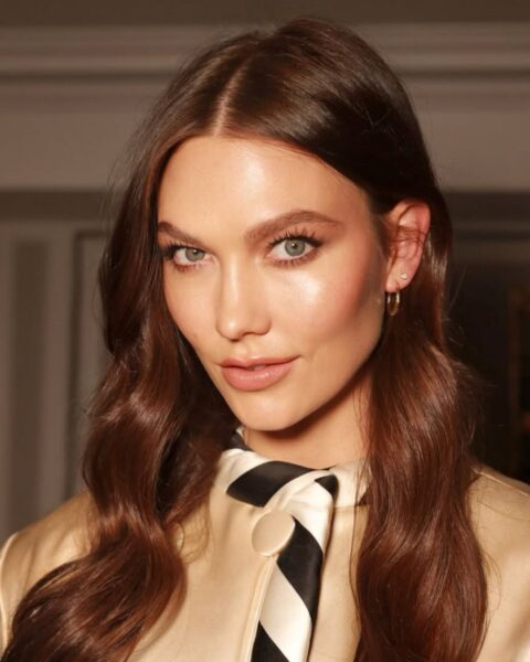 Portret Karlie Kloss z długimi, kasztanowymi włosami ułożonymi w fale. Ma promienny, świetlisty makijaż, podkreślający kości policzkowe i usta w kolorze nude. Ubrana jest w satynową, beżowo-złotą bluzkę z czarno-białym krawatem.