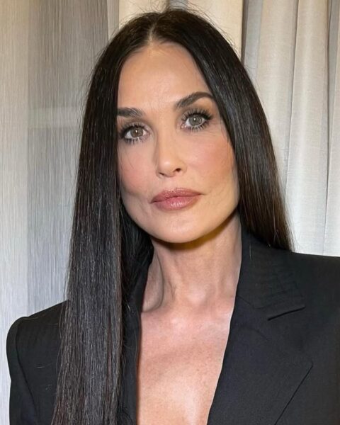 Portret Demi Moore w czarnej, eleganckiej marynarce z szerokimi ramionami i głębokim dekoltem. Ma długie, proste, ciemne włosy z przedziałkiem i subtelny makijaż z beżowymi ustami. Uosabia ponadczasową, klasyczną elegancję.