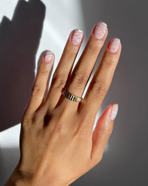 Nowoczesny, minimalistyczny manicure z artystycznym wzorem na naturalnej bazie. Kolor bazowy to delikatny nude w odcieniu milk-pink, a wzór to białe, falujące linie (styl abstract swirl lub wavy lines), które pokrywają całą płytkę paznokcia. Ten inspirujący i elegancki design na owalnych paznokciach jest idealny dla kobiet z klasą ceniących cichy luksus. Dłoń zdobi szeroki, fakturowany, złoty pierścionek w promieniach słonecznych.